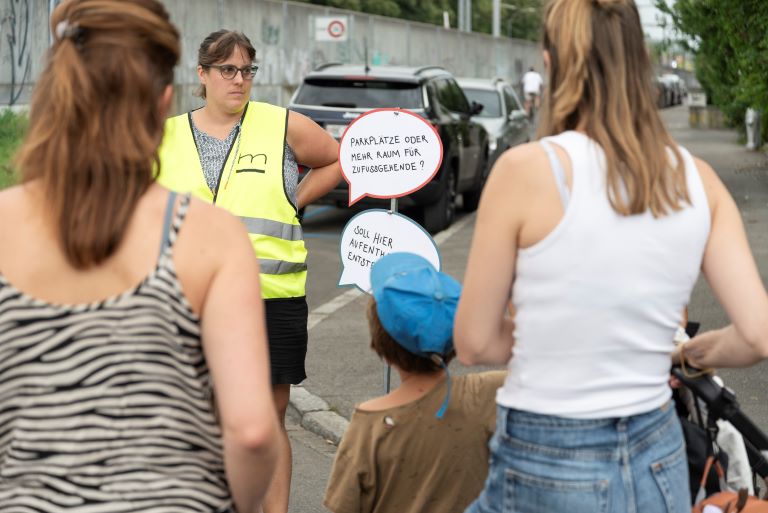 Quartierbevölkerung in Bern-Bümpliz betrachtet Sprechblasen zur Quartiergestaltung. Mitarbeiterin von Metron erklärt das Konzept.