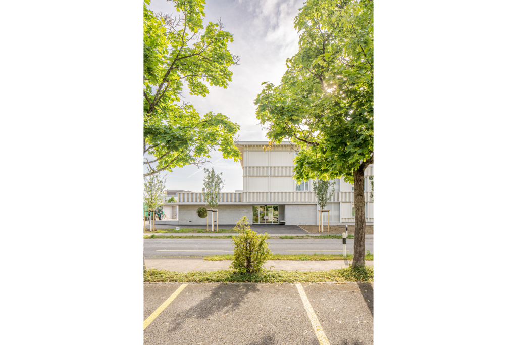 Blick über die Hauptstrasse zum Eingang. Das Volumen des Ateliers hebt sich als Betonfläche von der Holzverkleidung des übrigen Gebäudes ab.