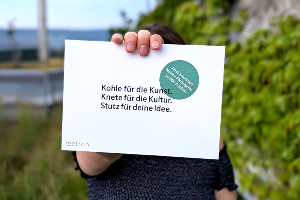 Foto. Eine Person hält den Metron-Förderpreis Flyer in die Kamera.