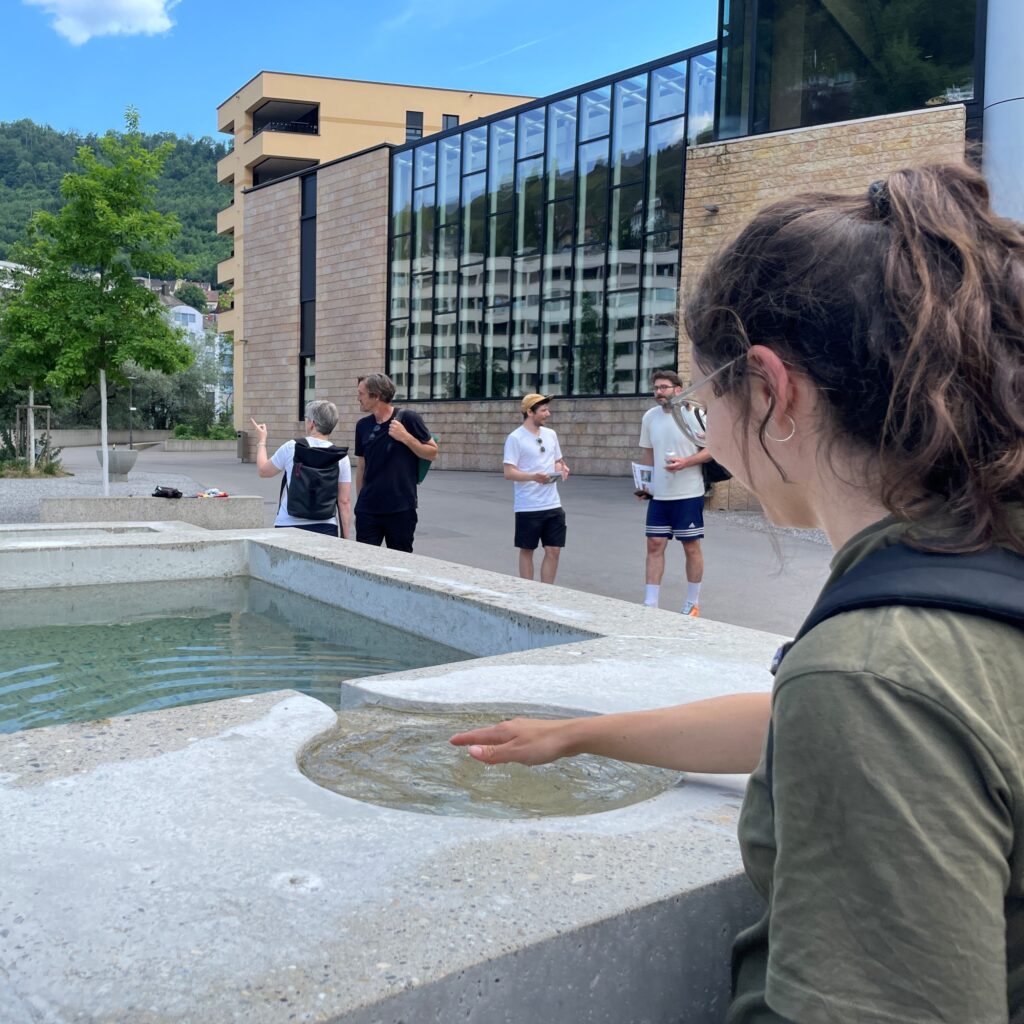 Bagni Popolari. Junge Frau hält ihre Hand in das Wasser von einem Brunnen. Im Hintergrund vier Personen.