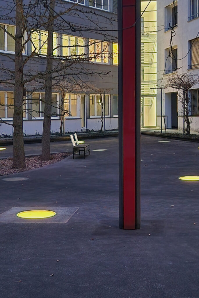 Hof eines Bürogebäudes mit Bank und Kunstwerk, aus der Tiefgarage scheint Licht durch runde Fenster im Asphalt